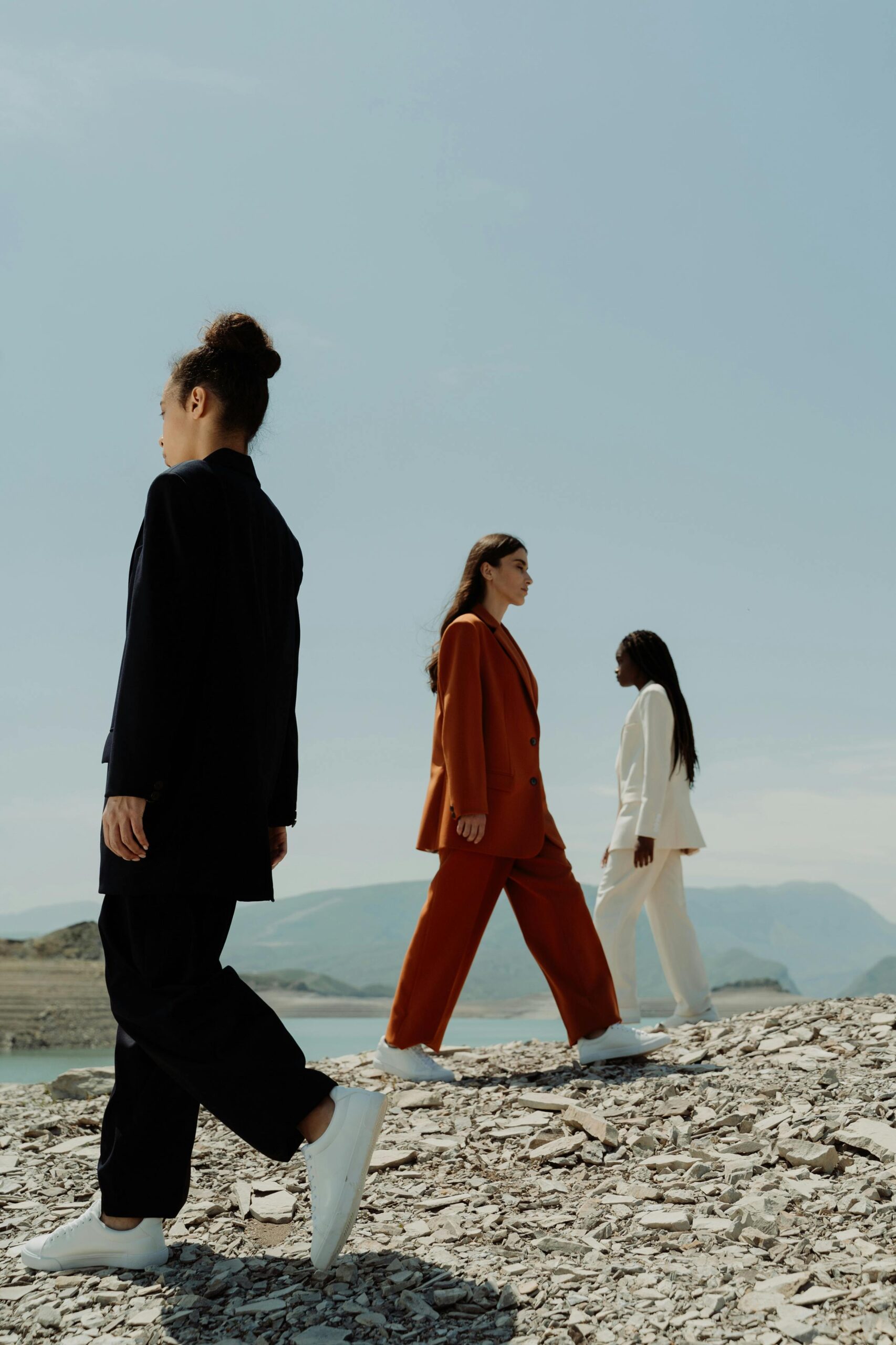 Three women in vibrant blazers walking on a rocky shore with a scenic backdrop.